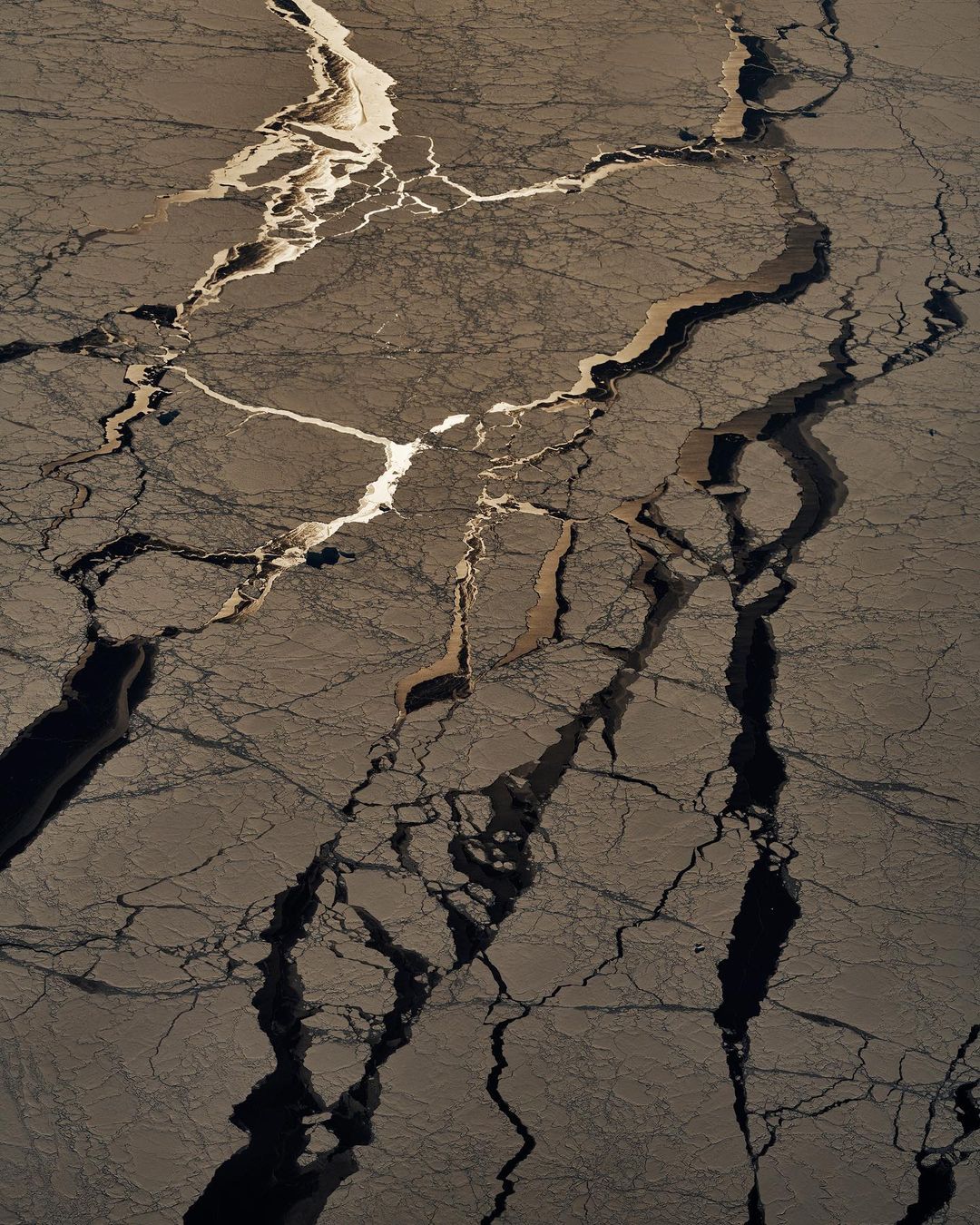 Aerial view of cracked ice formations with geometric patterns and contrasting light and dark tones in a natural environment.