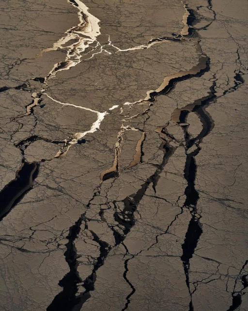 Aerial view of cracked ice formations with geometric patterns and contrasting light and dark tones in a natural environment.