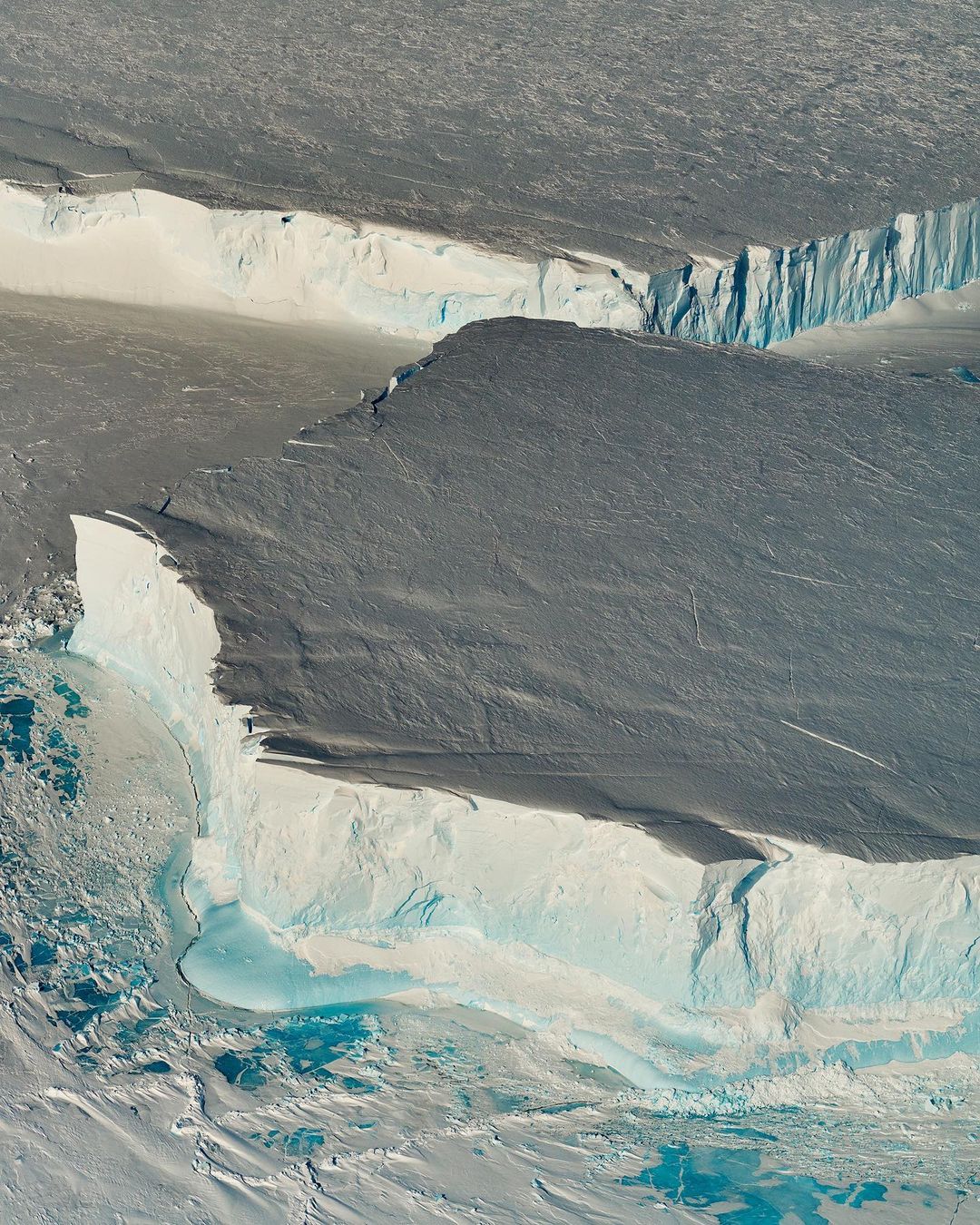 Aerial view of a large glacier showing layers of snow and ice, with a mix of blue, white, and grey coloring the natural landscape.