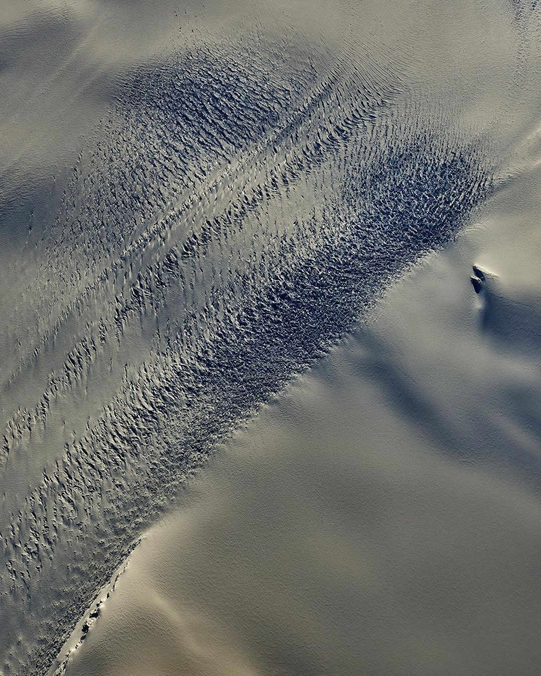 Aerial view captures stark beauty of snow-covered land, showcasing textured patterns and subtle gradations of white and grey in natural light.