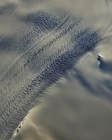 Aerial view captures stark beauty of snow-covered land, showcasing textured patterns and subtle gradations of white and grey in natural light.
