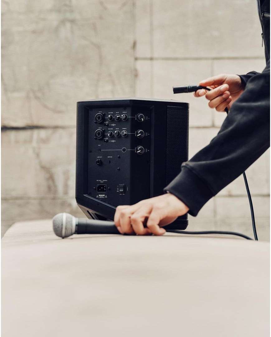 Black speaker on a beige surface with a hand connecting a microphone cable, set against a neutral wall background. Music equipment.