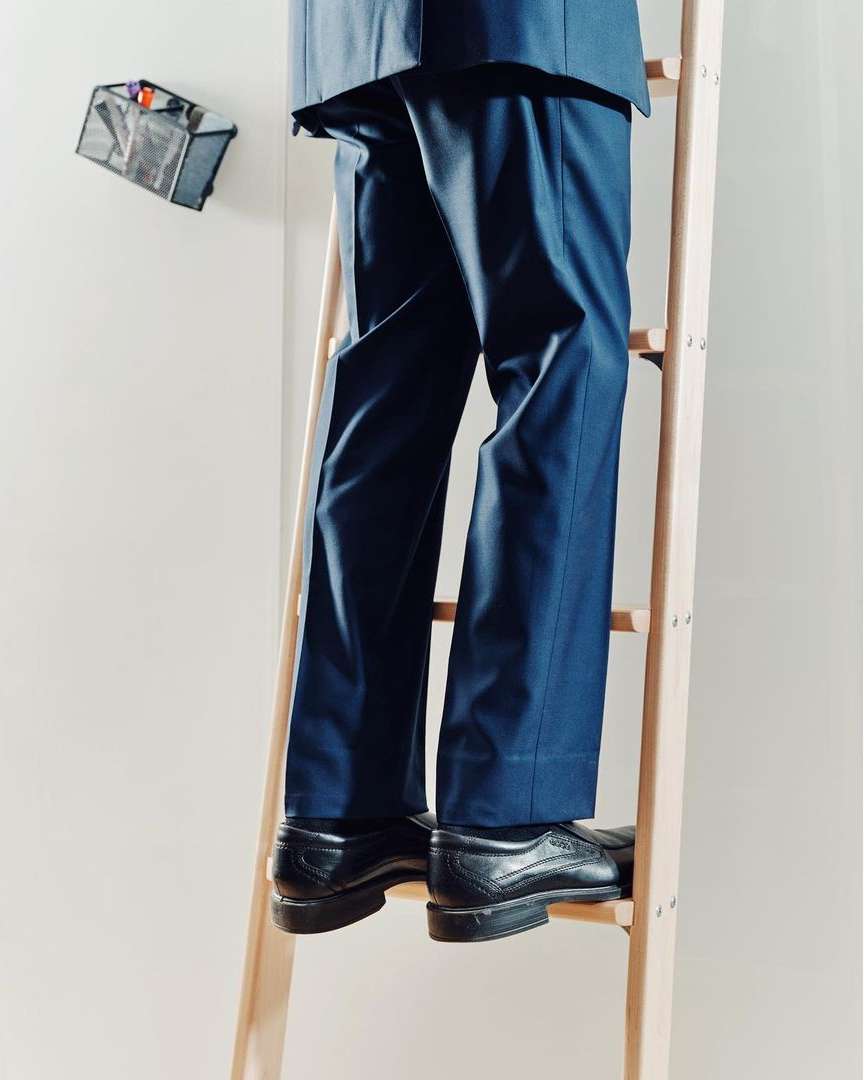 Torso shot of a person in a suit climbing a wooden ladder indoors; a tool bag hangs on the wall, creating a sense of anticipation or home repair.