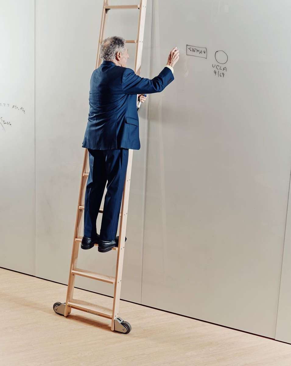 Man in a suit on a rolling ladder writes on a whiteboard. He is indoors with white walls and light wood floor.