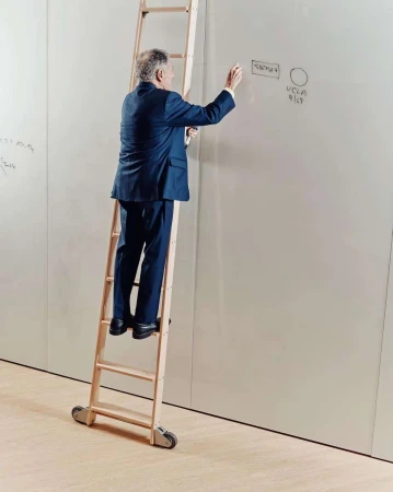 Man in a suit on a rolling ladder writes on a whiteboard. He is indoors with white walls and light wood floor.