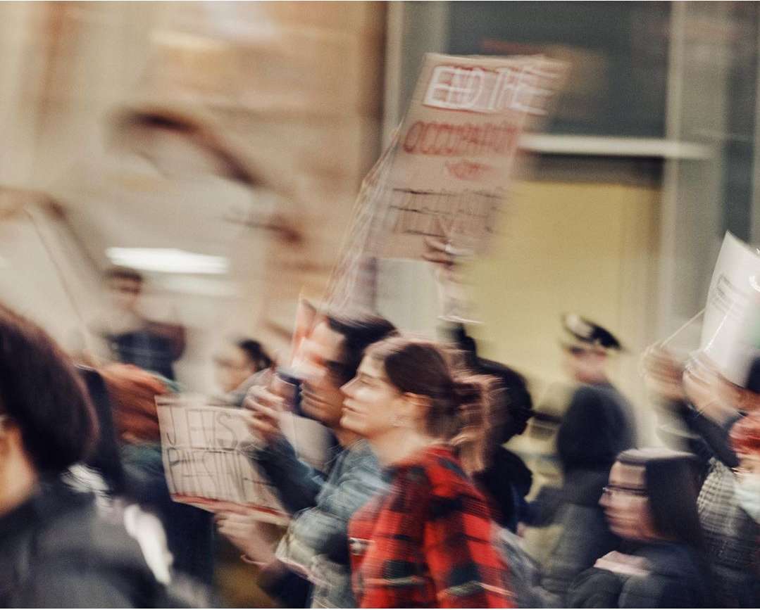 People march holding signs in a blurred outdoor scene. A woman in a red plaid shirt is visible in focus amongst the crowd.