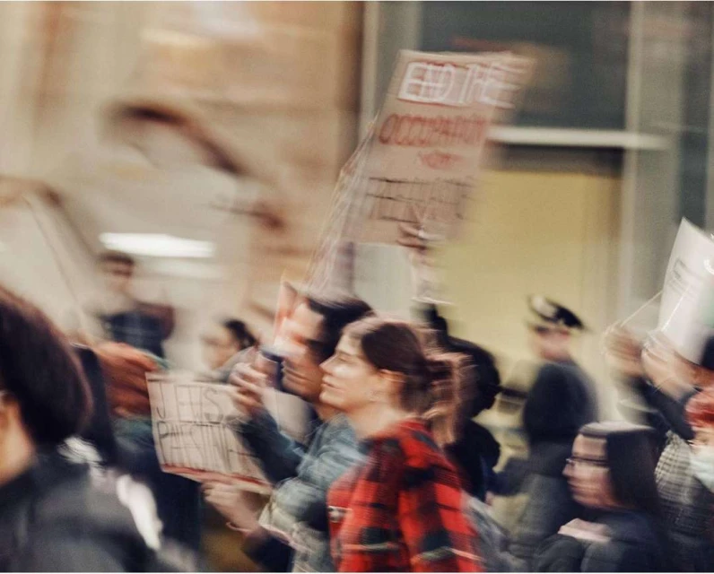 People march holding signs in a blurred outdoor scene. A woman in a red plaid shirt is visible in focus amongst the crowd.