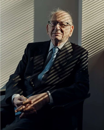 Elderly man in a dark suit and glasses seated indoors with striped window shadow patterns across him, photographed by Thomas Prior.