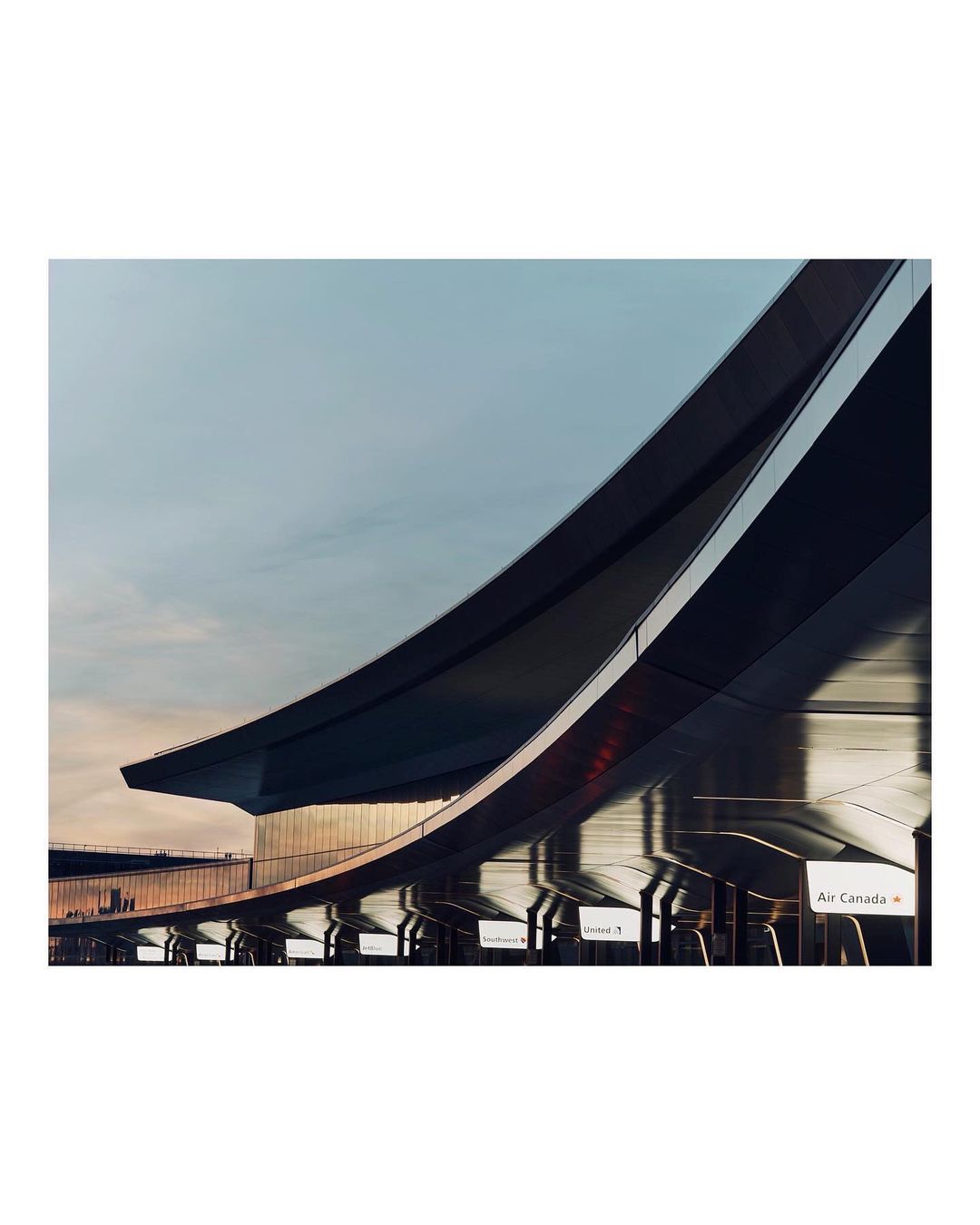 Exterior of an airport with curved architecture, featuring layered rooflines and signage, set against a pastel sky at daytime.