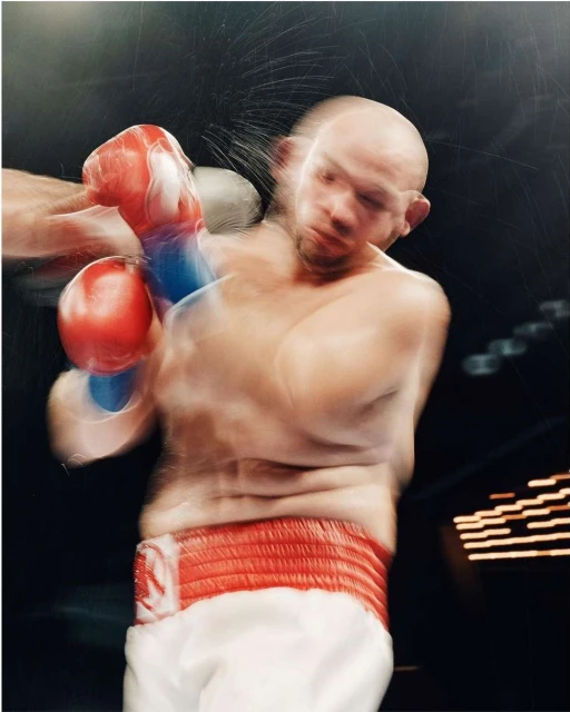 Close-up action shot of a bald adult male boxer in motion wearing red boxing gloves and a red waistband, in an indoor boxing arena.