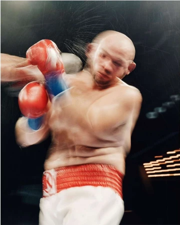Close-up action shot of a bald adult male boxer in motion wearing red boxing gloves and a red waistband, in an indoor boxing arena.