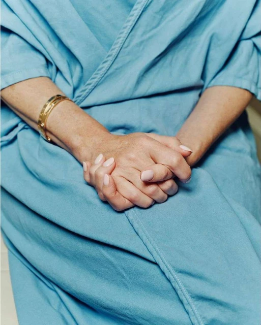 Close-up of a woman's folded hands wearing a blue hospital gown and gold bangle, seated in a bright indoor environment.