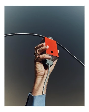 Close-up of an adult hand holding a red electronic component attached to grey and black cables against a grey background.