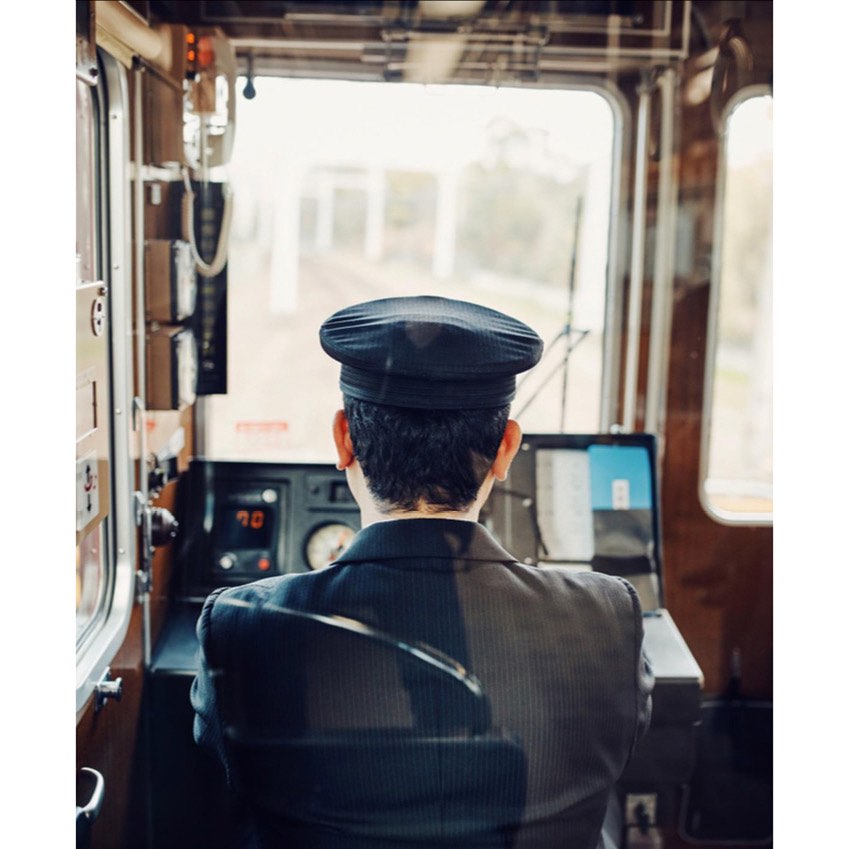 Train engineer in uniform operates vintage train from inside the cabin, seen from behind, with exterior visible through windshield.