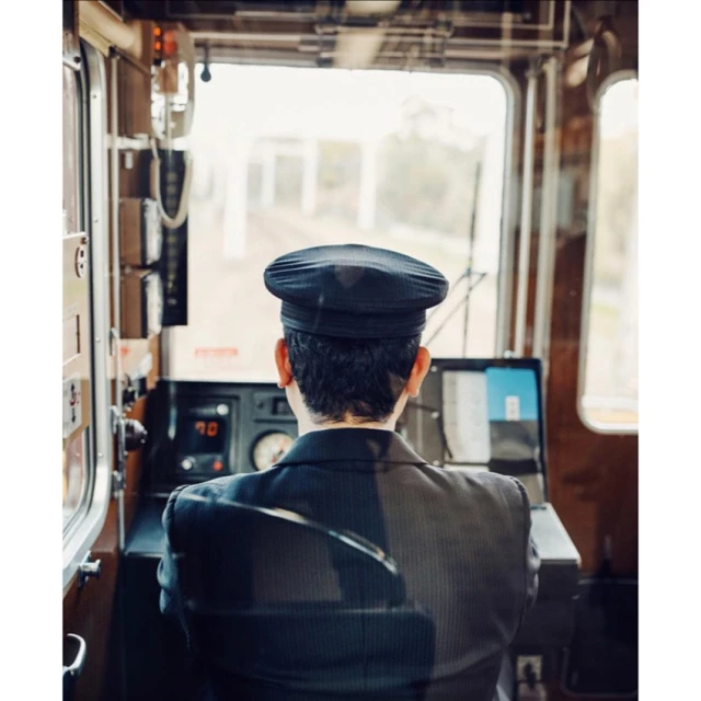 Train engineer in uniform operates vintage train from inside the cabin, seen from behind, with exterior visible through windshield.