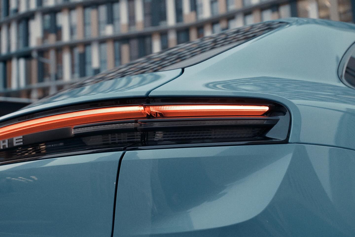 Close-up of the rear light bar on a blue Porsche. The backdrop features an out-of-focus building with many windows.
