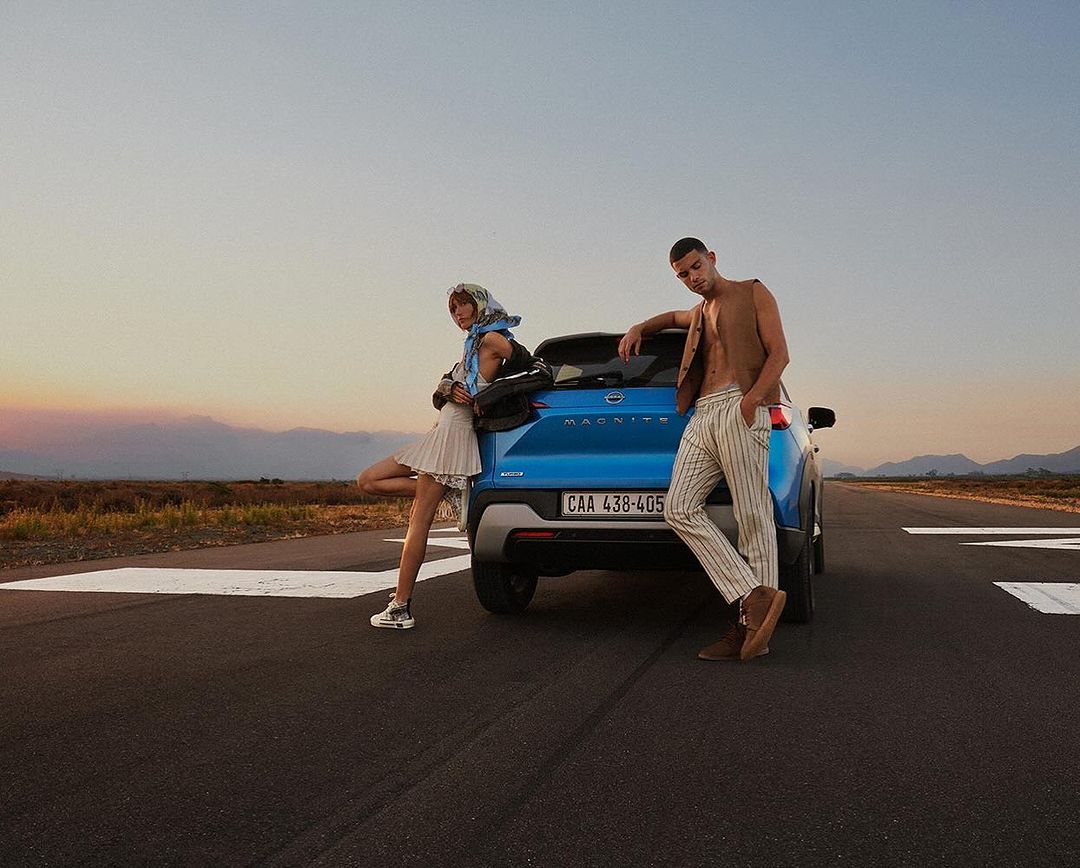 Two fashion models pose with a blue Nissan Magnite on an airfield runway at sunset. Mountains in the background.