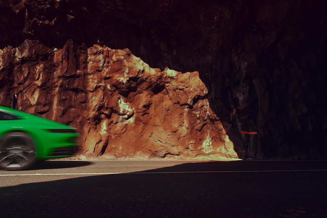 Motion blurred shot of a green sports car driving on an asphalt road passing a craggy rock formation in a dimly lit outdoor environment.