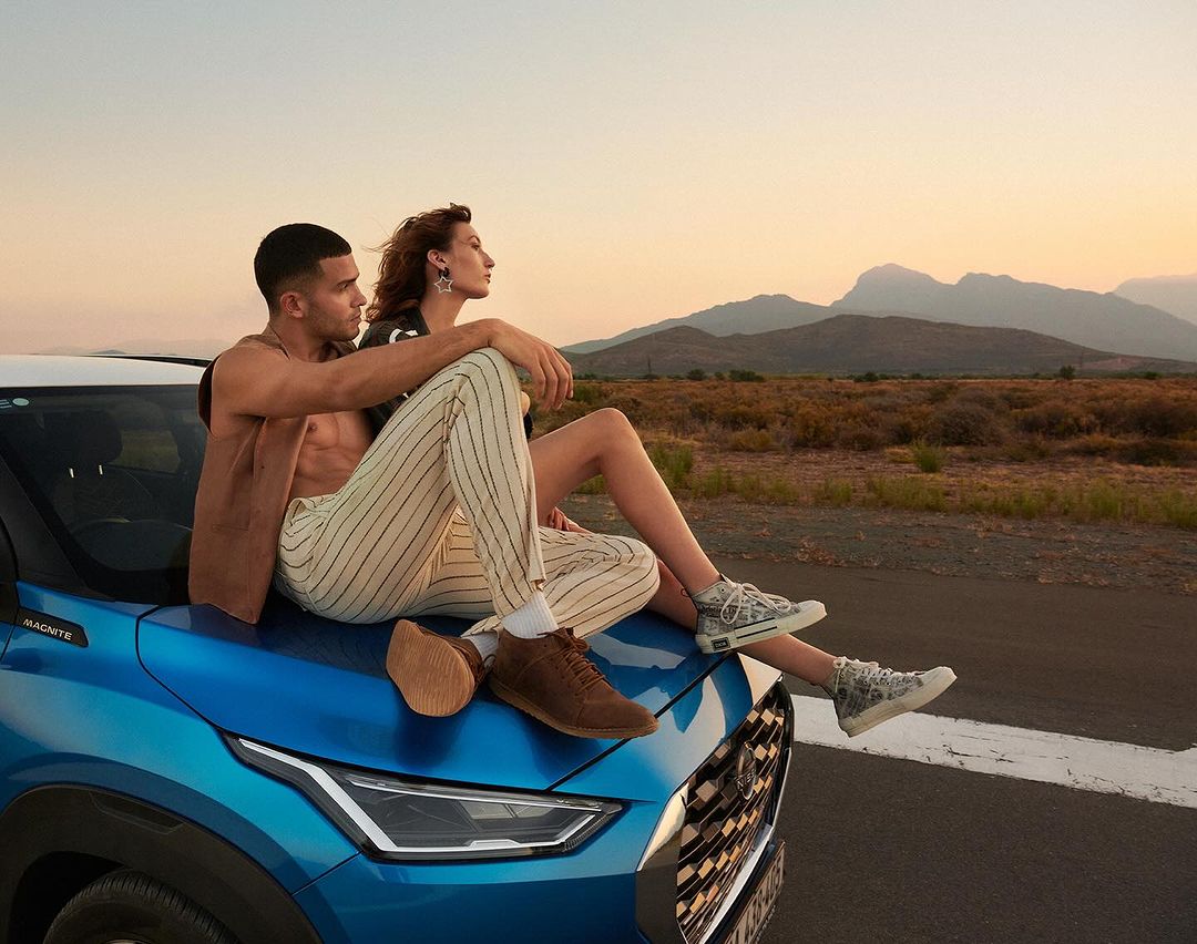 A couple sits on a blue car parked along a road near mountains in an outdoor setting, featuring a man with a bare chest and tan pants.