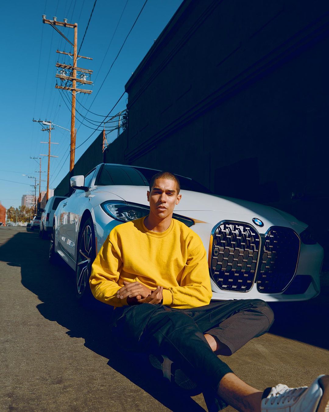 A young adult male in a yellow sweatshirt sits on an asphalt street with a modern white BMW car on a sunny day in an urban setting.