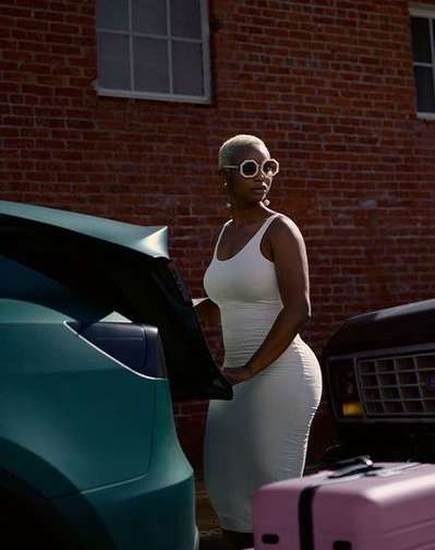 Adult woman in a white dress opening the trunk of a teal car outdoors on a sunny day with a brick building in the background.
