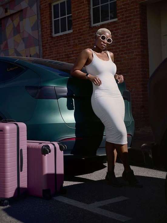 Stylish woman in white dress posing next to a green car with two pink suitcases in an urban setting on a sunny day with shadows.
