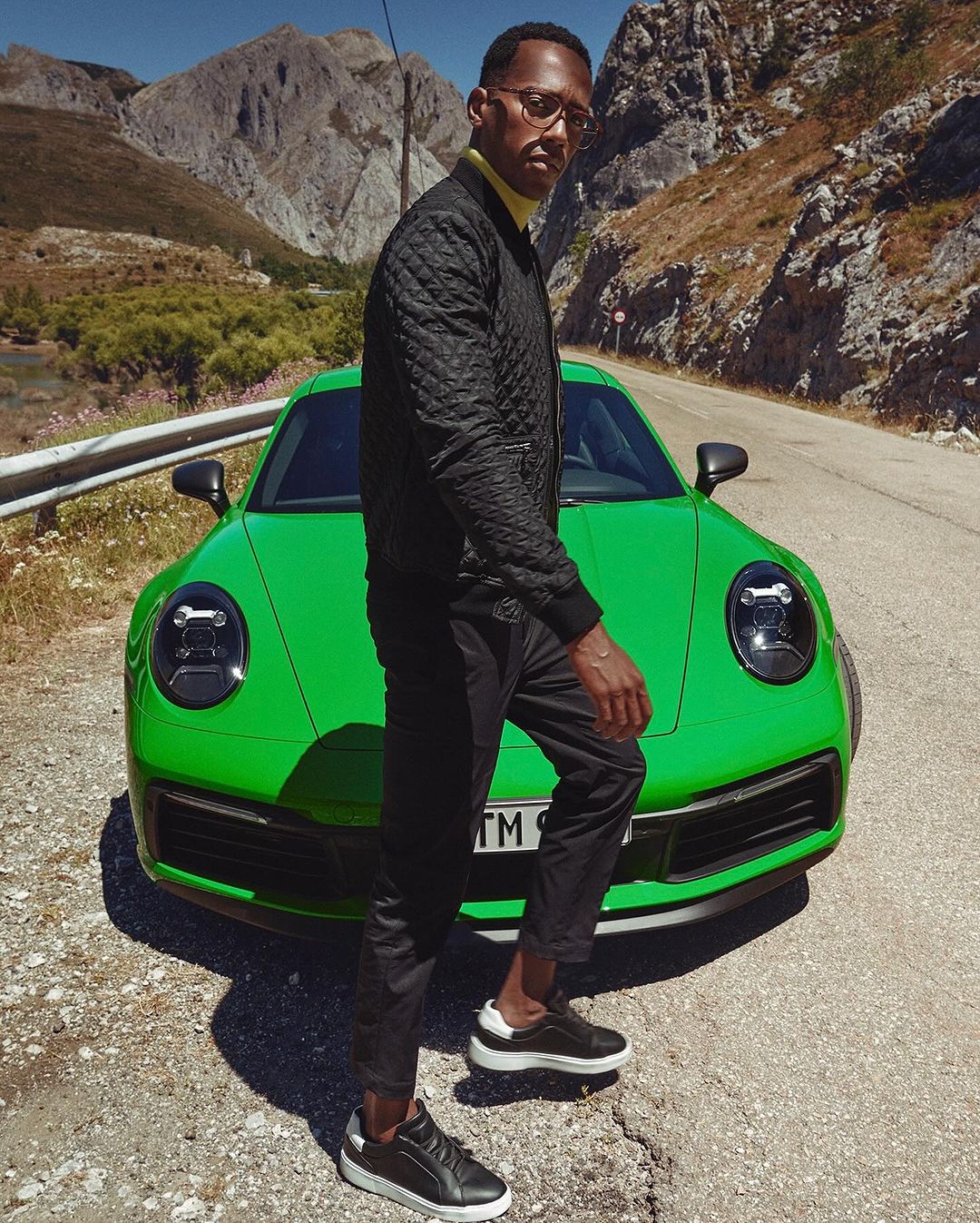 Adult man stands next to bright green sports car in a mountain landscape wearing black quilted jacket, pants, and stylish sneakers.