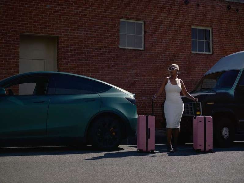 A woman in a white dress stands outside next to an electric car and van, holding the handles of two pink rolling suitcases in an urban setting.