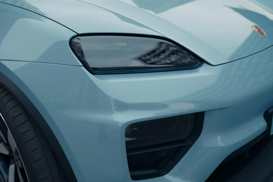 Close-up of a light-blue Porsche, showcasing its front headlight and sleek design. The car reflects building windows.