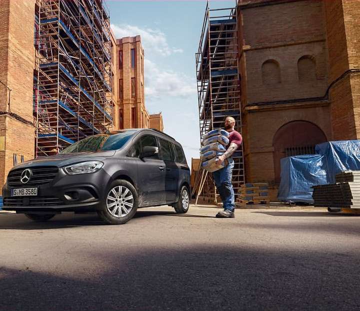 Dark grey Mercedes Citan passenger van parked on street with an adult male carrying stack of cement bags in front of construction site.