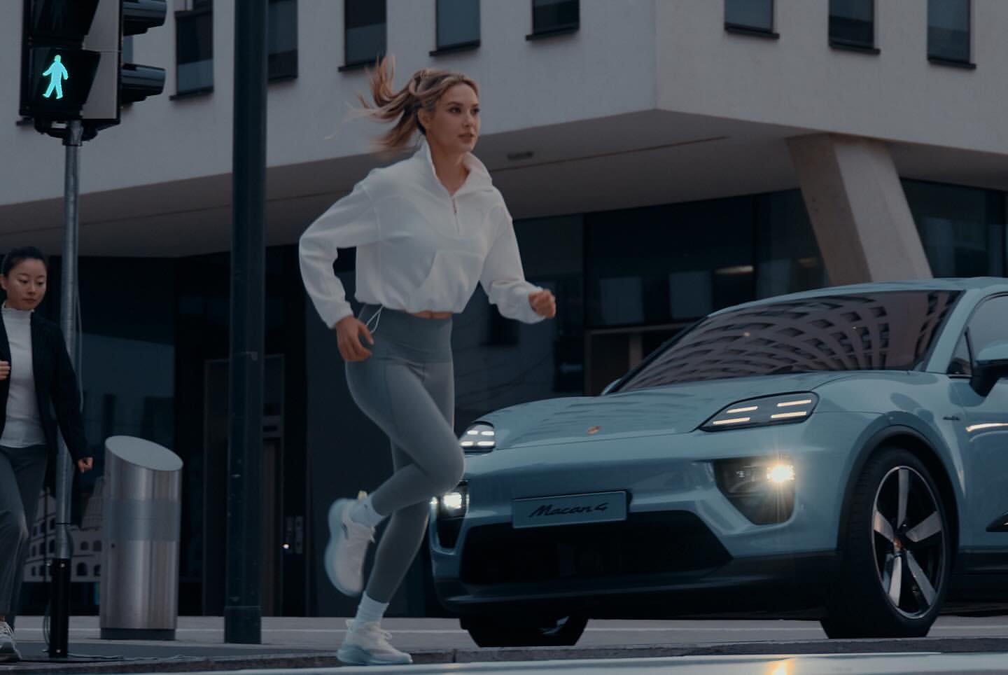Blond woman in athletic wear runs across an urban crosswalk near a light blue Porsche Macan 4 with a building in the background.