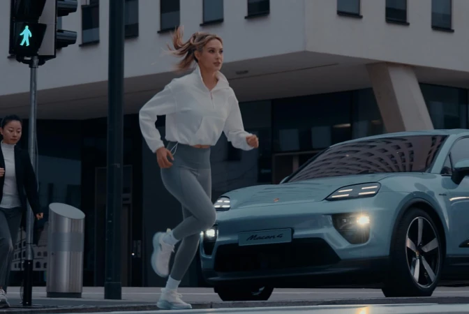 Blond woman in athletic wear runs across an urban crosswalk near a light blue Porsche Macan 4 with a building in the background.