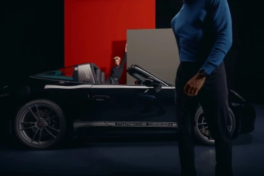 A man in a blue sweater stands in the foreground with a black sports car and a woman leaning against a panel in the studio.