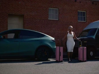 Woman in white dress standing between a dark car and van with pink luggage against brick wall