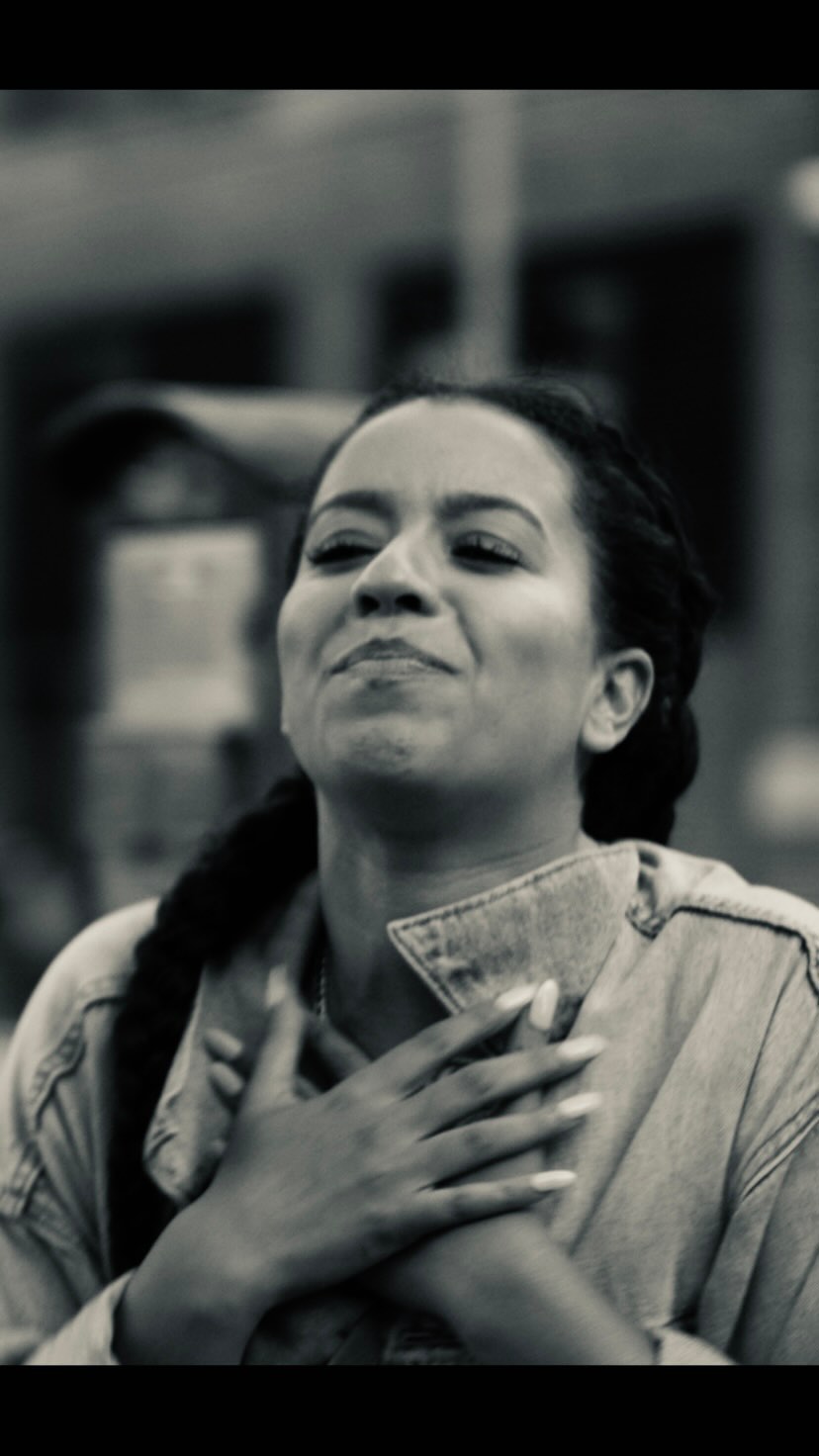 Monochromatic portrait of a woman with a braid wearing a light denim jacket and crossing her hands over her chest in an outdoor setting.