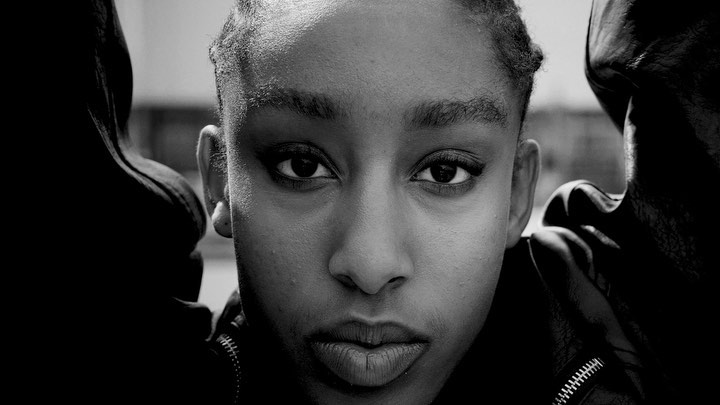Close-up portrait of an adult woman with short hair and dark skin, wearing a black leather jacket, in a black-and-white outdoor shot.