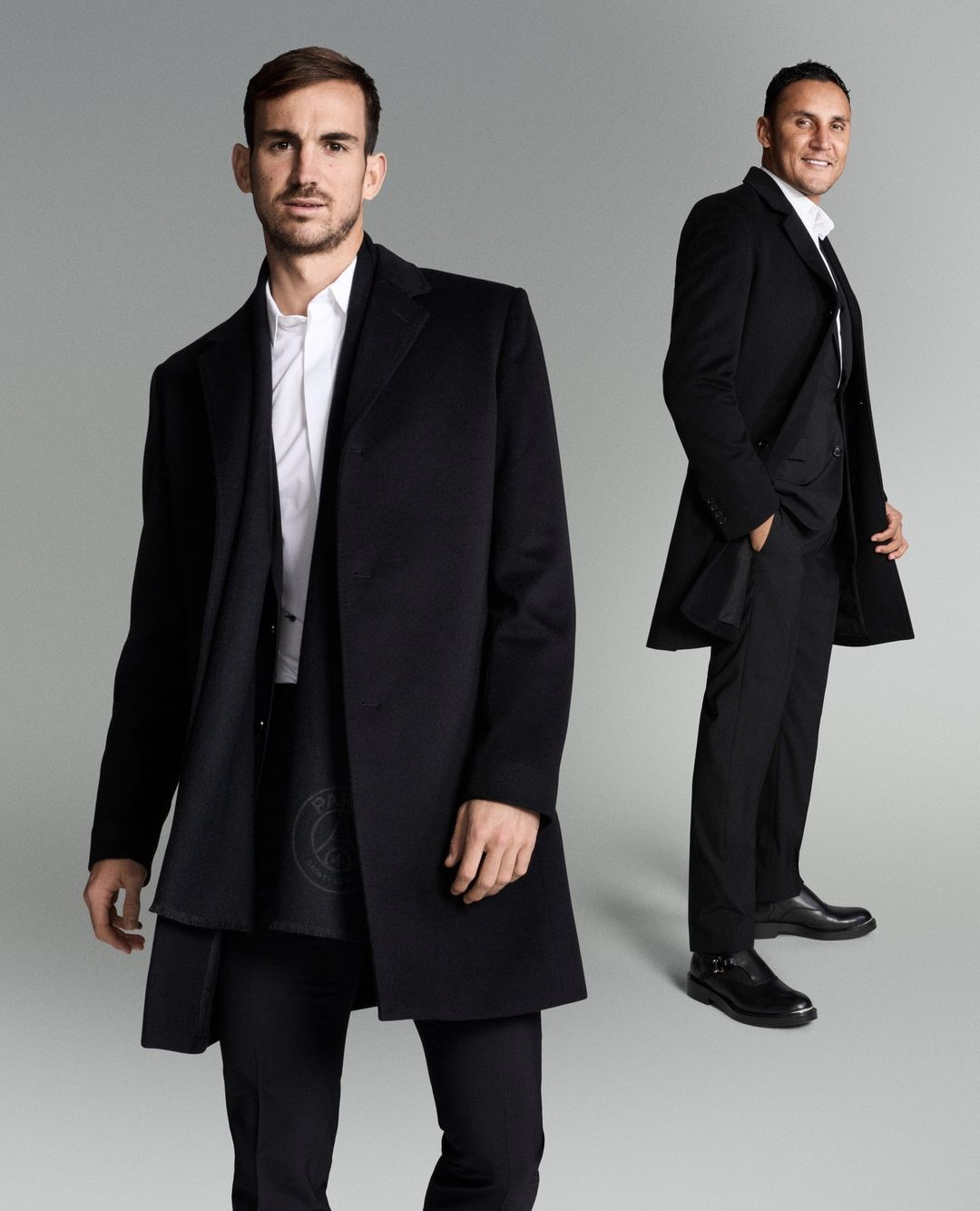 Two adult men in black overcoats and suits pose in a studio setting against a gradient gray background in a medium studio shot.