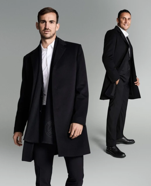 Two adult men in black overcoats and suits pose in a studio setting against a gradient gray background in a medium studio shot.