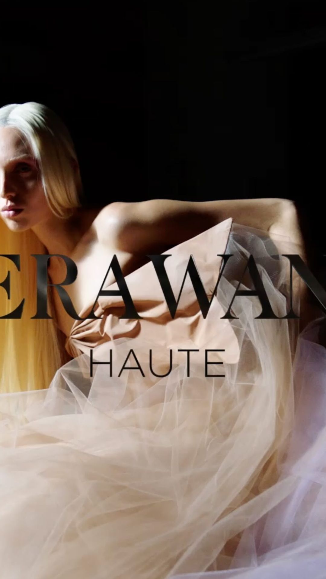 Adult female fashion model in a light tan dress with soft tulle fabric and blond hair in a studio setting with dark background.