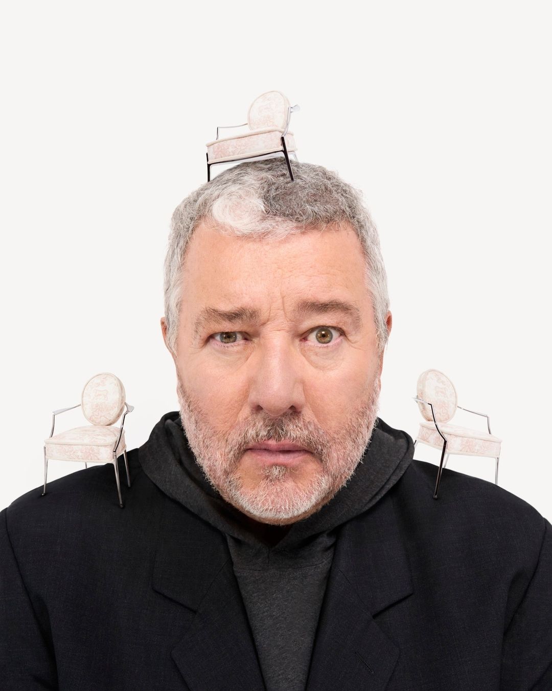 A man with grey hair poses, wearing a black coat, with miniature chairs balanced on his head and shoulders against a white backdrop.