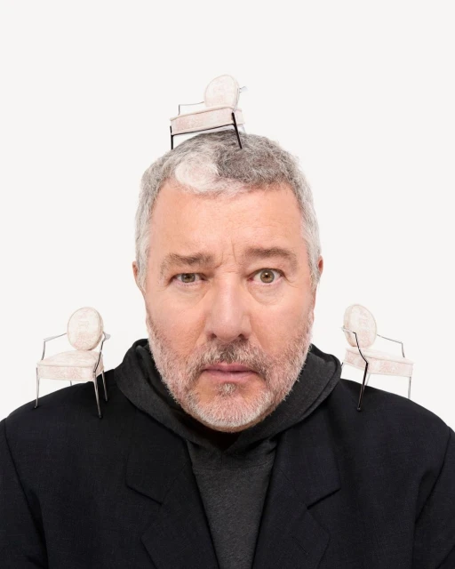 A man with grey hair poses, wearing a black coat, with miniature chairs balanced on his head and shoulders against a white backdrop.