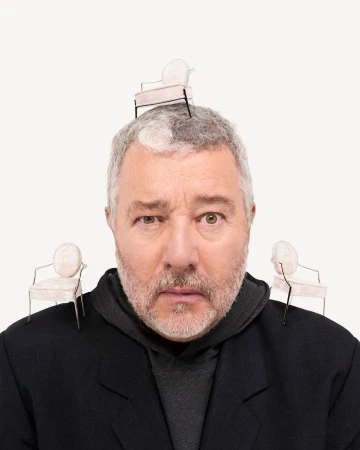 A man with grey hair poses, wearing a black coat, with miniature chairs balanced on his head and shoulders against a white backdrop.