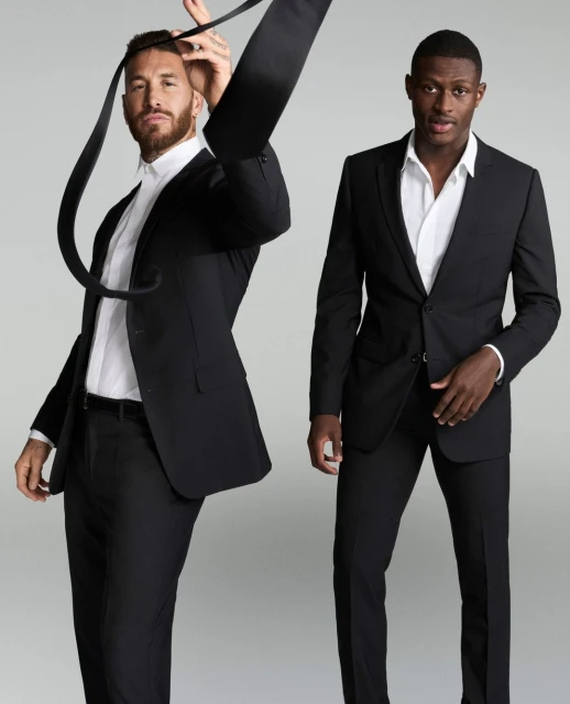 Two adult men in black suits pose in a studio against a neutral background, one holding a necktie above his head in a promotional fashion shot.