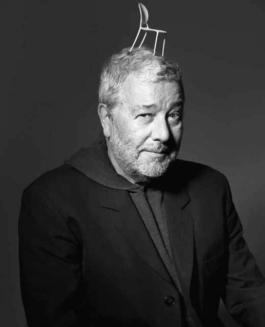 Black and white portrait of an adult man with short gray hair and a beard, wearing a suit jacket, with a miniature chair balanced on his head.