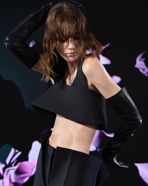 Woman in black crop top and skirt poses with long black gloves against a floral background in a fashion shot with moody lighting.