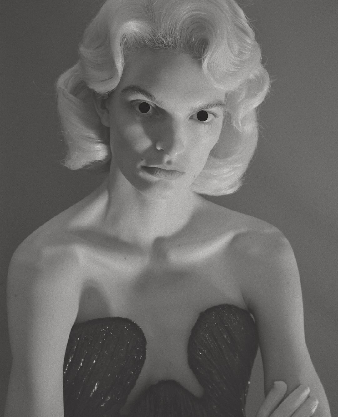 Portrait of a model with sculpted blonde hair and dark voids for eyes, wearing a dark strapless dress, by photographer Till Janz.