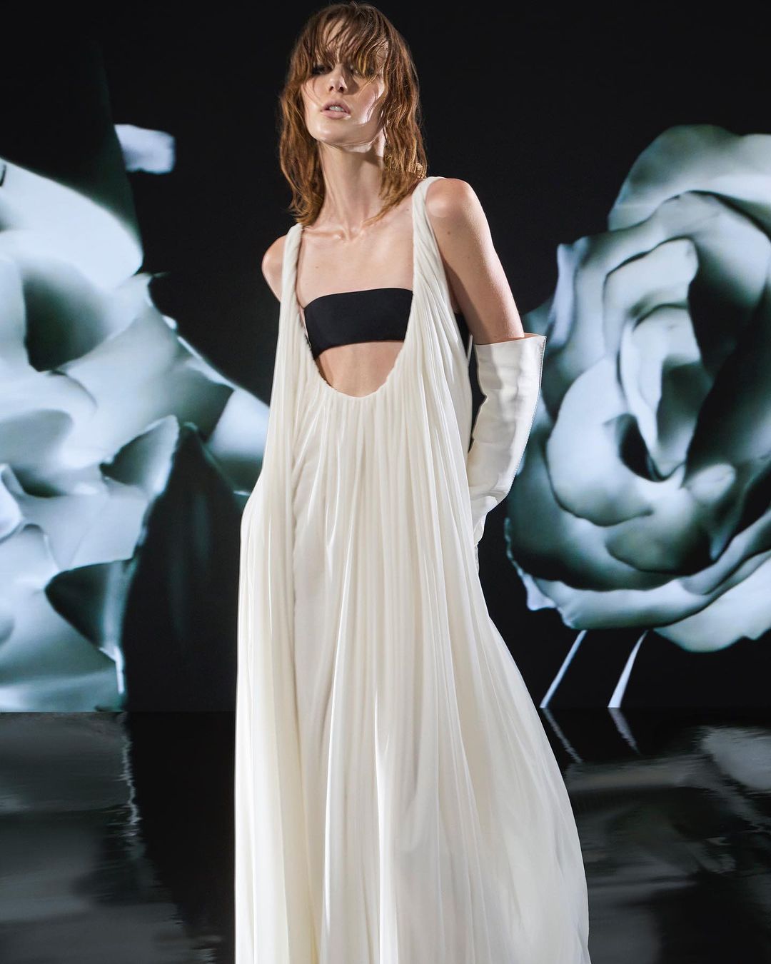 Fashion shoot featuring a slender adult female wearing a white dress and opera gloves, posing in front of a floral pattern backdrop.
