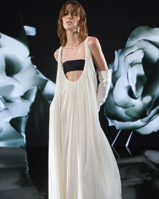 Fashion shoot featuring a slender adult female wearing a white dress and opera gloves, posing in front of a floral pattern backdrop.