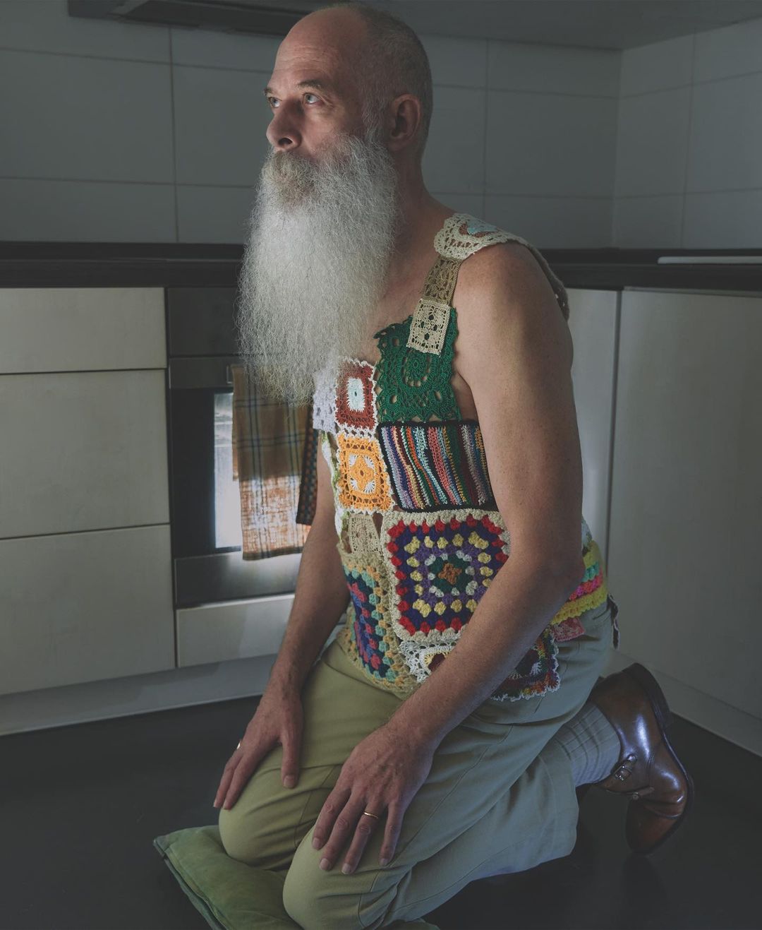 Senior man kneeling indoors on a green cushion, wearing a patchwork crochet vest and khaki pants in a kitchen setting with an oven.