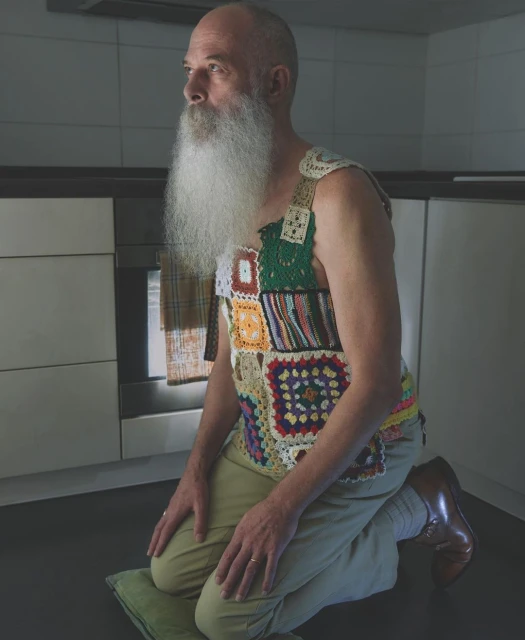 Senior man kneeling indoors on a green cushion, wearing a patchwork crochet vest and khaki pants in a kitchen setting with an oven.